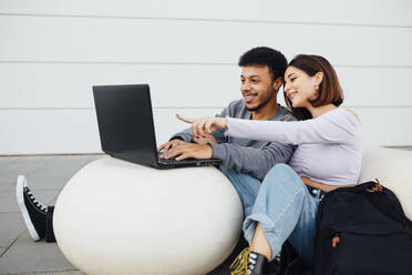 Lächelndes Paar sitzt und benutzt Laptop auf weißem Betonball - MIMFF00304