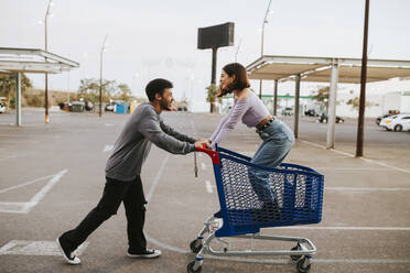 Boyfriend pushing girlfriend standing in shopping cart on road - MIMFF00309