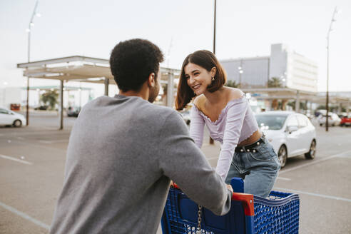 Young man pushing woman standing in shopping cart on road - MIMFF00310