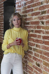 Fashionable mature woman holding tea cup while leaning on brick wall in kitchen - VEGF03252