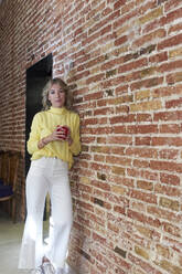 Mature woman with tea cup leaning on brick wall in kitchen - VEGF03253