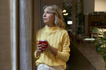 Modische reife Frau schaut durch das Fenster und hält eine Teetasse in der Küche - VEGF03258