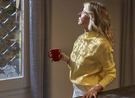 Ältere Frau schaut durch ein Fenster und hält eine Teetasse - VEGF03260