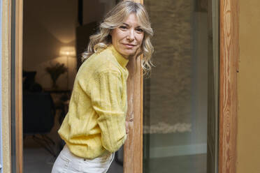 Lächelnde reife Frau mit blondem Haar lehnt sich an die Tür zu Hause - VEGF03262