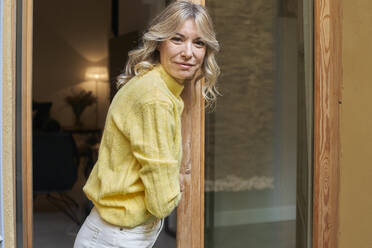 Smiling mature woman with blond hair leaning on door at home - VEGF03262