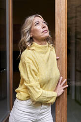 Nachdenkliche reife Frau im gelben Pullover, die aufschaut, während sie sich zu Hause an die Tür lehnt - VEGF03264