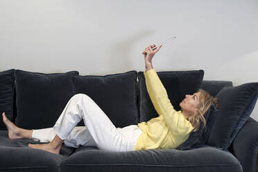 Reife Frau mit erhobenen Händen, die ein digitales Tablet benutzt, während sie zu Hause auf dem Sofa liegt - VEGF03274