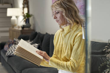 Ältere Frau im gelben Pullover liest ein Buch auf dem Sofa zu Hause - VEGF03275