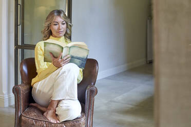 Smiling woman reading book while sitting on armchair at home - VEGF03276