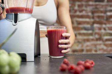 Ältere Frau hält ein Glas mit Himbeer-Smoothie an einem Mixer auf einer Kücheninsel - VEGF03301