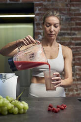 Reife Frau gießt Himbeer-Frucht-Smoothie in Glas in der Küche - VEGF03302