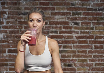 Mature woman drinking fruit smoothie against brick wall in kitchen - VEGF03304