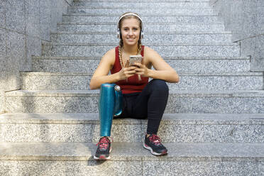 Sportlerin mit Beinprothese lächelt, während sie auf einer Treppe sitzend ein Mobiltelefon benutzt - IFRF00156