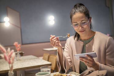 Lächelnde Geschäftsfrau, die etwas isst und dabei ihr Handy benutzt, in einem modernen Cafe - SDAHF01016