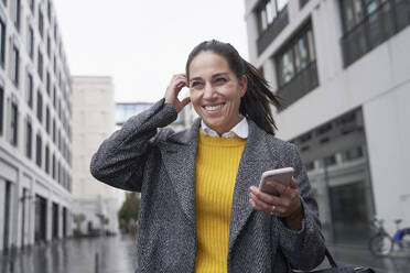 Smiling businesswoman with hand in hair using mobile phone while standing on street - SDAHF01029