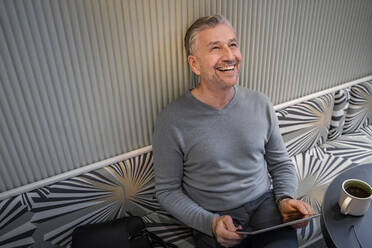 Happy businessman with digital tablet looking up while sitting at modern cafe - SDAHF01045