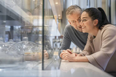 Lächelnde Geschäftsfrau, die die Kuchenauslage betrachtet, während sie neben einem Mann in einem modernen Cafe steht - SDAHF01048