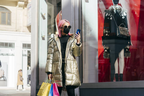 Modische Frau mit Gesichtsmaske, die Einkaufstüten trägt, während sie ein Mobiltelefon benutzt und am Schaufenster steht - MARF00025