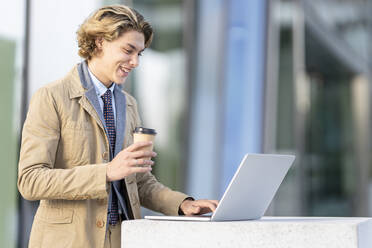 Lächelnder Geschäftsmann mit Kaffeetasse, der im Freien stehend am Laptop arbeitet - GGGF00419