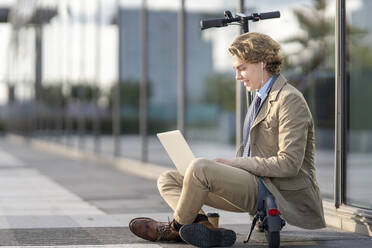 Young businessman with in-ear headphones using laptop while sitting on electric push scooter - GGGF00426