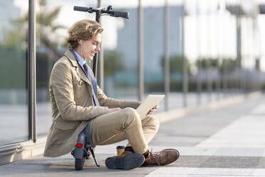 Businessman wearing in-ear headphones using laptop while sitting on electric push scooter - GGGF00427