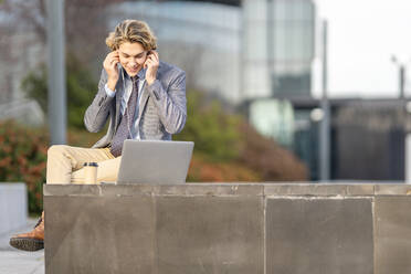 Geschäftsmann passt In-Ear-Kopfhörer an, während er im Freien sitzend am Laptop arbeitet - GGGF00430