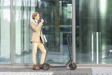 Businessman with laptop drinking coffee while standing by electric push scooter against glass wall - GGGF00436