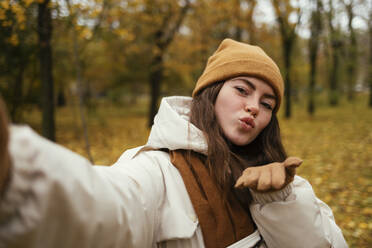 Beautiful young woman blowing kiss while taking selfie in autumn park - OYF00264