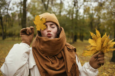 Young woman with eyes closed puckering while covering eye with autumn leaf in public park - OYF00270