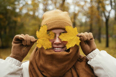 Happy young woman wrapped in scarf covering eyes with autumn leaf in public park - OYF00273