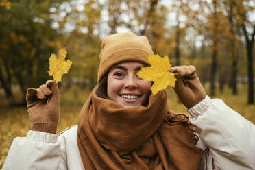 Happy young woman covering eye with autumn leaf in public park - OYF00275