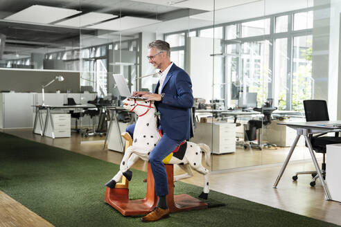 Glücklicher männlicher Unternehmer, der am Laptop arbeitet, während er auf einem Schaukelpferd am Arbeitsplatz sitzt - PESF02363