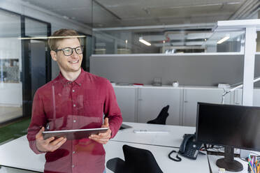 Happy businessman looking away while holding digital tablet at work place - PESF02382