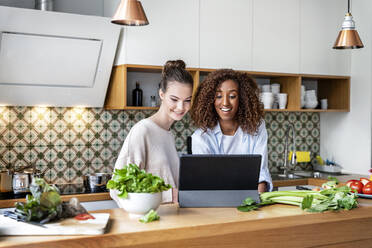 Glückliche weibliche Mitarbeiter, die sich in der Küche am Arbeitsplatz ein Rezept auf einem Tablet ansehen - PESF02397