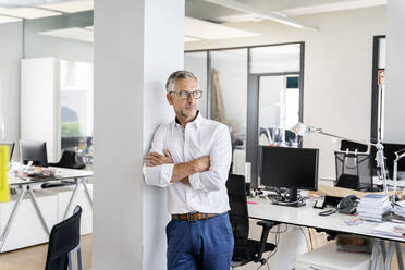 Confident businessman with arms crossed leaning on column in office - PESF02418