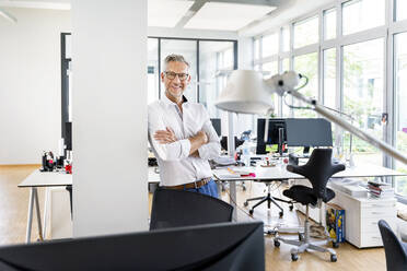 Smiling male entrepreneur with arms crossed leaning on column at work place - PESF02419