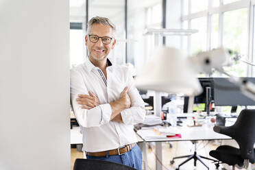 Happy male professional with arms crossed leaning on wall in office - PESF02420