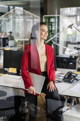Lächelnde Geschäftsfrau mit Laptop, die wegschaut, während sie sich auf den Schreibtisch im Büro stützt - PESF02431