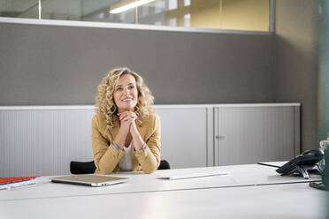 Smiling female professional with hands clasped day dreaming at desk in office - PESF02444