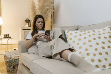 Young woman using mobile phone while relaxing on sofa at home - MTBF00775