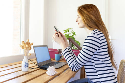 Rothaarige Frau mit Laptop, die ein Mobiltelefon benutzt, während sie zu Hause sitzt - AFVF07779