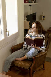 Woman with book drinking coffee while sitting on chair at home - AFVF07783