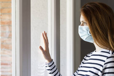 Redhead woman wearing protective face mask standing by window at home during COVID-19 - AFVF07785