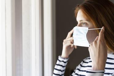 Young woman wearing protective face mask while standing at home during COVID-19 - AFVF07786
