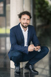 Male entrepreneur crouching with mobile phone at office lobby - JOSEF02558