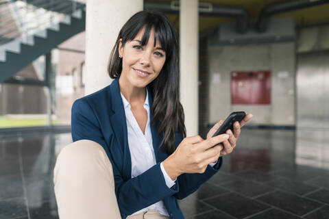 Lächelnde Geschäftsfrau hält ihr Smartphone in der Hand, während sie in der Büro-Lobby sitzt, lizenzfreies Stockfoto
