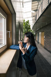 Businesswoman with smart phone looking through window while standing in corridor of office - JOSEF02570