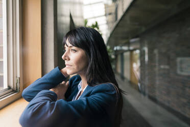 Weibliche Unternehmerin lehnt sich an, während sie durch ein Fenster im Büro schaut - JOSEF02571