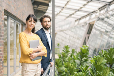 Professional male and female colleagues with laptop standing by railing in office corridor - JOSEF02623