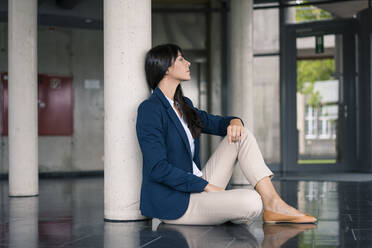 Businesswoman contemplating while sitting by column in office lobby - JOSEF02650